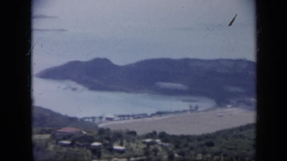1959 Venezuela 8mm frame with a wide elevated view of a sheltered bay, sandy shoreline, dark wooded hills, and small distant buildings.