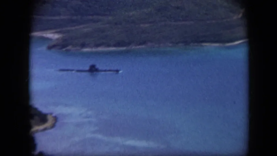 1959 Venezuela 8mm frame with a long low vessel moving across calm blue water below a green shoreline.
