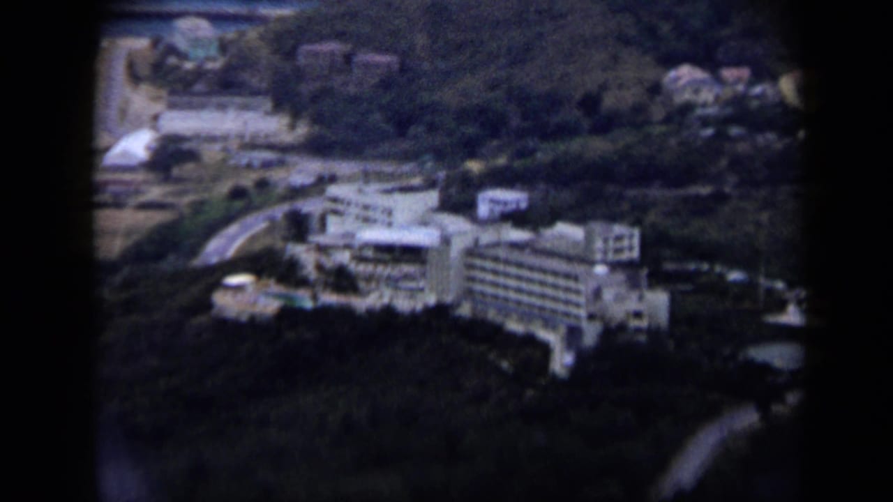 1959 Venezuela 8mm frame showing a large white hotel-like complex, winding roads, pools, and surrounding hills.