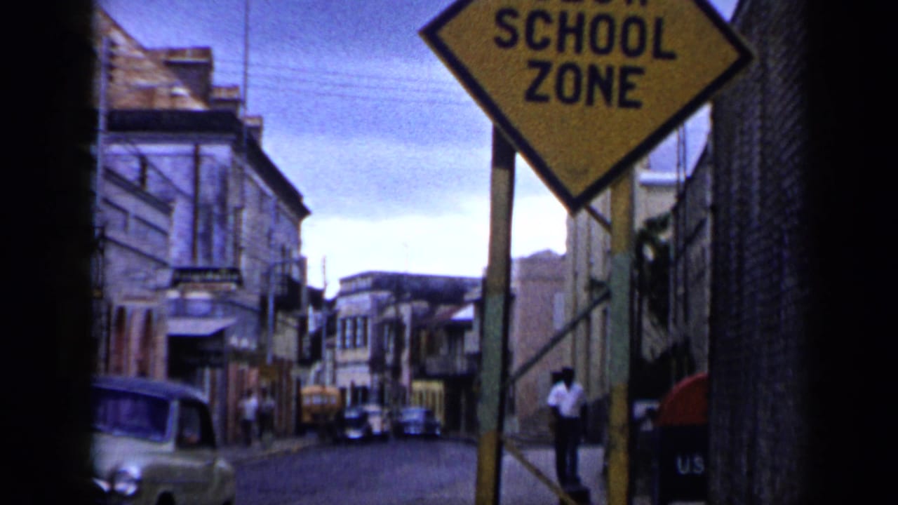 1959 Venezuela street view with a yellow school zone sign, older cars, buildings, and people in the distance.