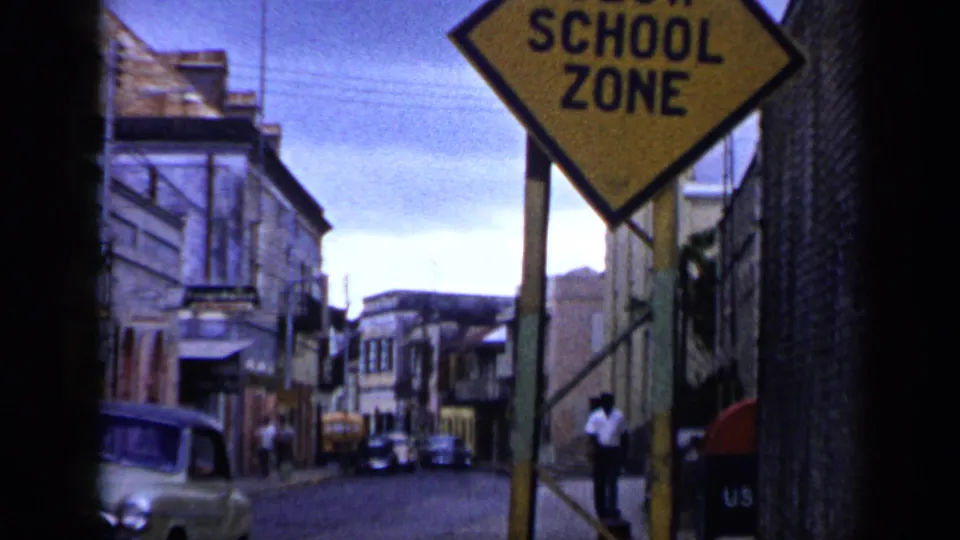 1959 Venezuela street view with a yellow school zone sign, older cars, buildings, and people in the distance.