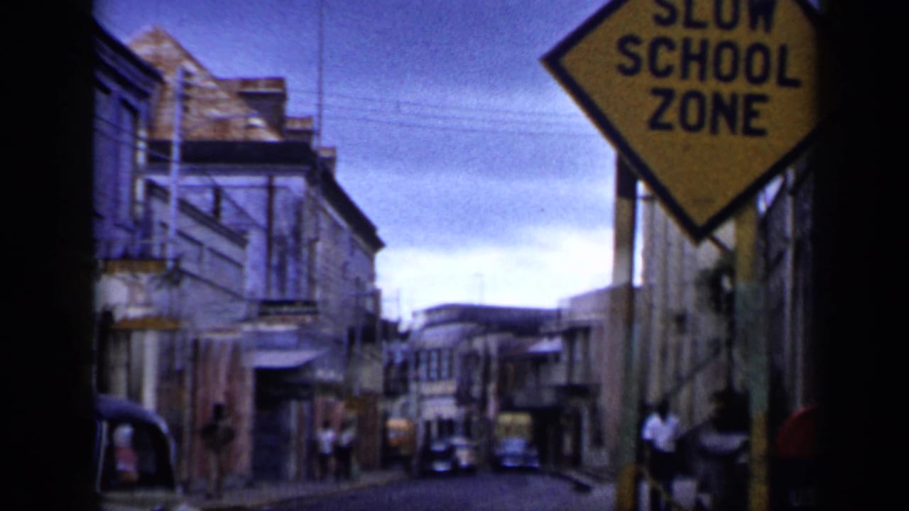 1959 Venezuela 8mm frame showing a yellow school-zone sign above a narrow tropical street with pedestrians, parked cars, and weathered buildings.