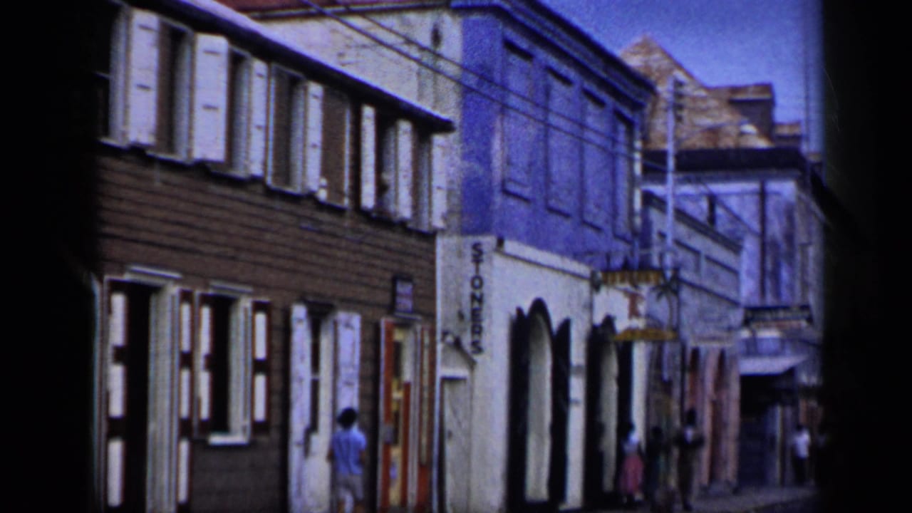 Street-front buildings with shuttered windows, a vertical sign, pedestrians, and utility wires.