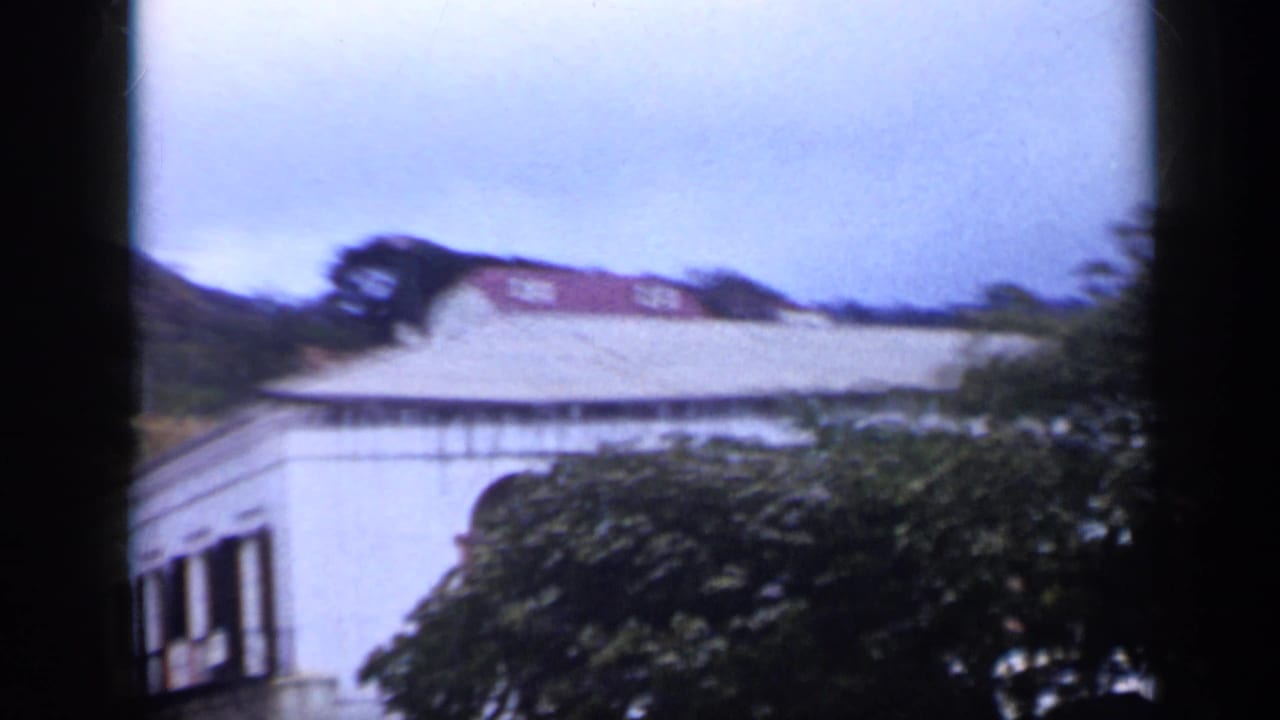 1959 Venezuela 8mm view of a white building and roofline partly covered by trees against hillside terrain.