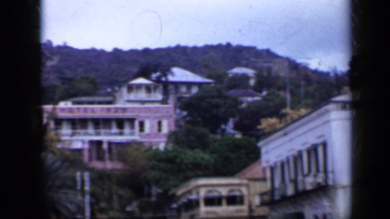 1959 Venezuela frame showing a pastel hillside building, white neighboring structures, trees, and sloped terrain.