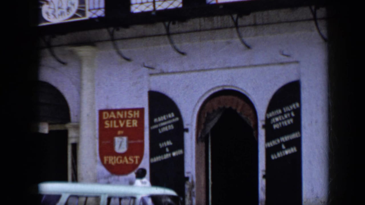 1959 Venezuela 8mm frame showing a hotel frontage with arched entry, Danish Silver by Frigast sign, dark shutters, and a light car at the curb.