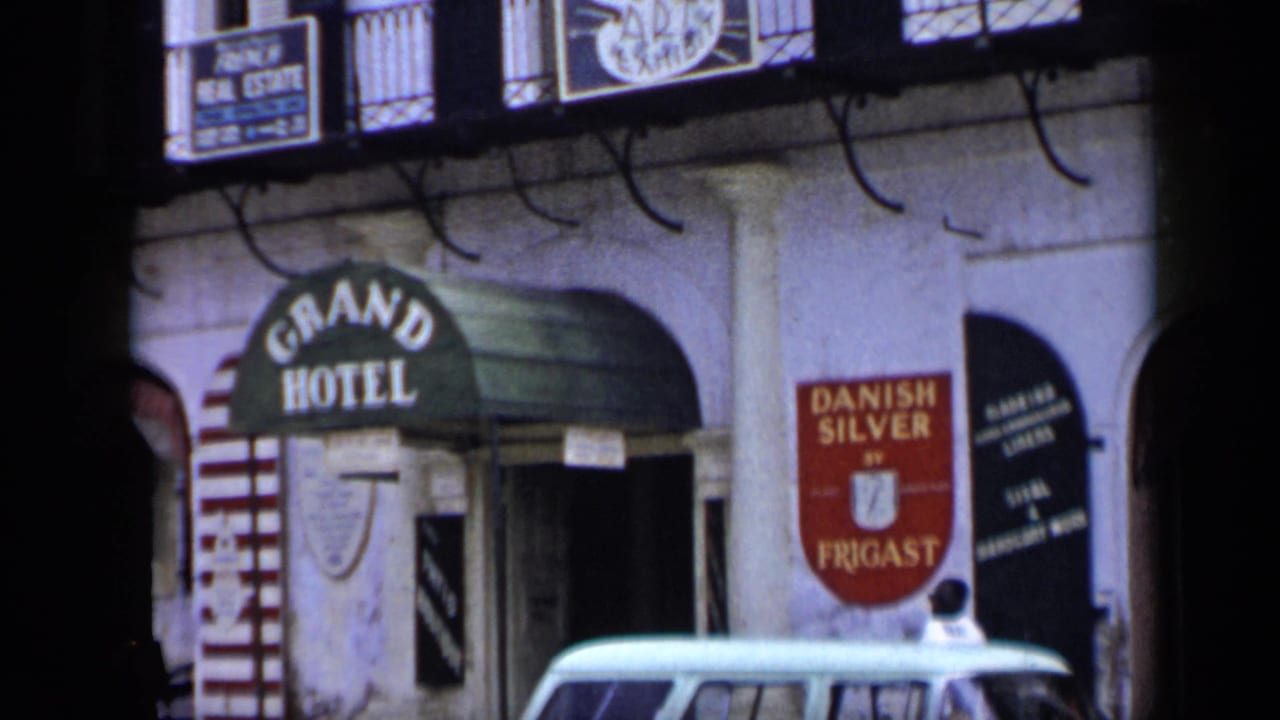 1959 Venezuela 8mm frame centered on a green Grand Hotel awning beside a red Danish Silver by Frigast sign and a parked light car.
