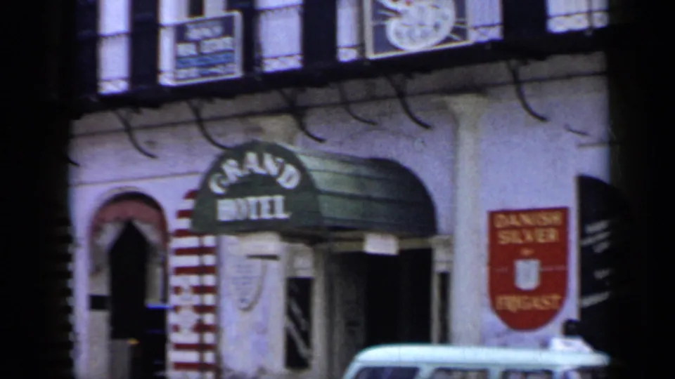 1959 Venezuela 8mm frame with angled handheld view of the Grand Hotel awning, adjacent signage, arched entry, and light-colored car.