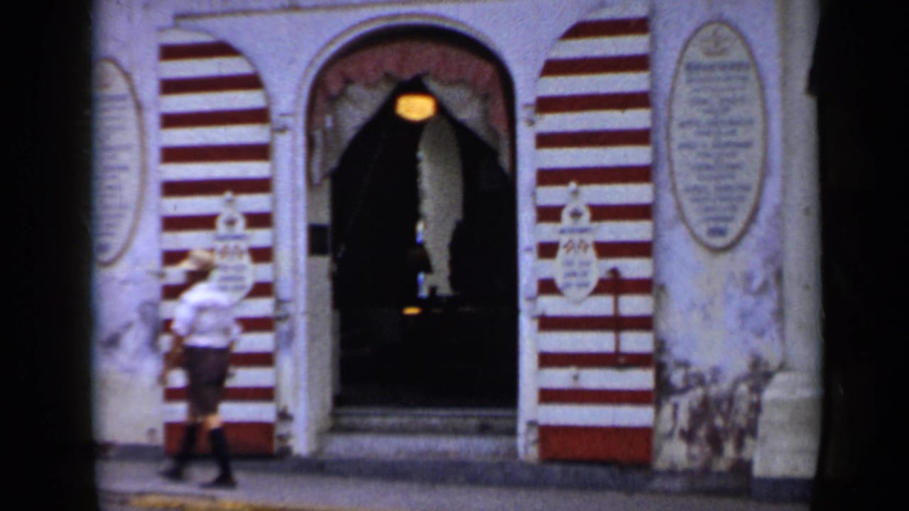1959 Venezuela 8mm frame showing a red-and-white striped Grand Hotel entrance as a man in a light shirt and hat walks by.