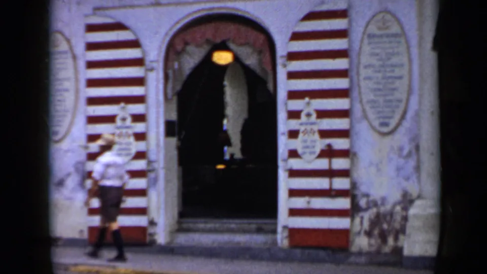 1959 Venezuela 8mm frame showing a red-and-white striped Grand Hotel entrance as a man in a light shirt and hat walks by.