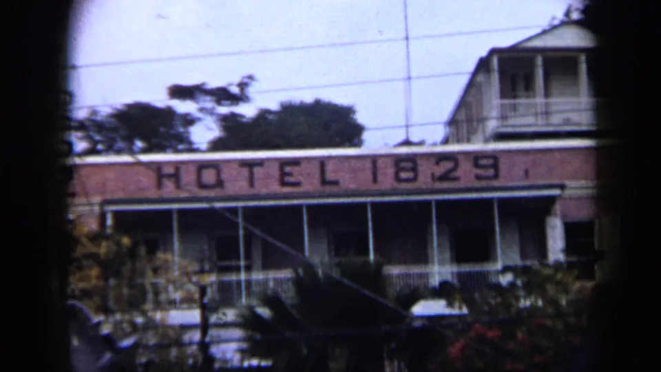 1959 Venezuela home-movie frame showing the Hotel 1829 sign, upper balcony, palm foliage, and overhead utility lines.
