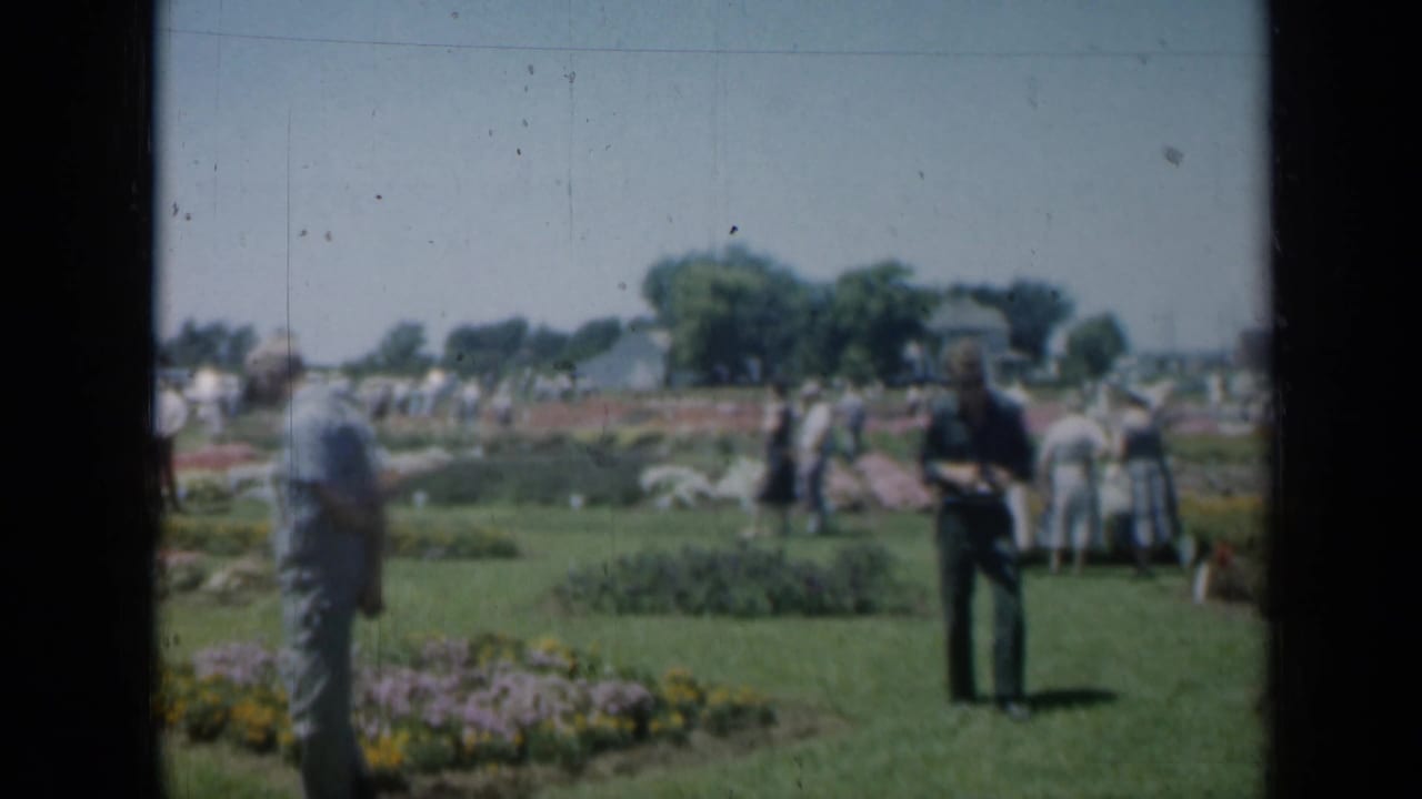 Adults in period clothing appear in a Missouri garden, with cars and trees visible beyond the flower beds.