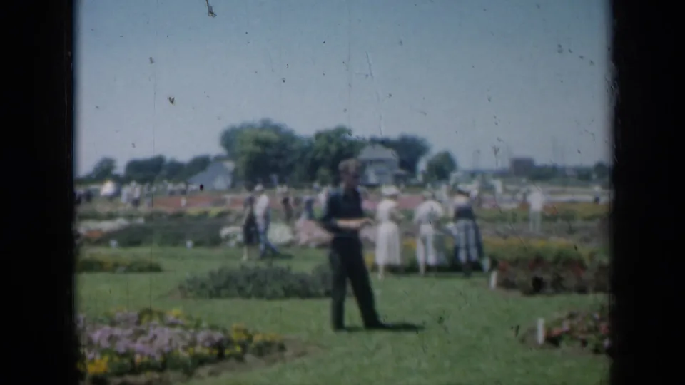 1961 Missouri home-movie frame with a man crossing the grass and several adults near colorful garden plantings.