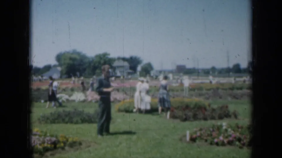 Soft 8mm frame of adults meandering through a Missouri garden with flower beds, lawn, cars, and trees in view.