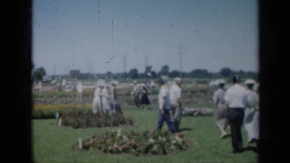Missouri 1961 garden scene with adults walking past flower beds, white markers, and distant utility poles.
