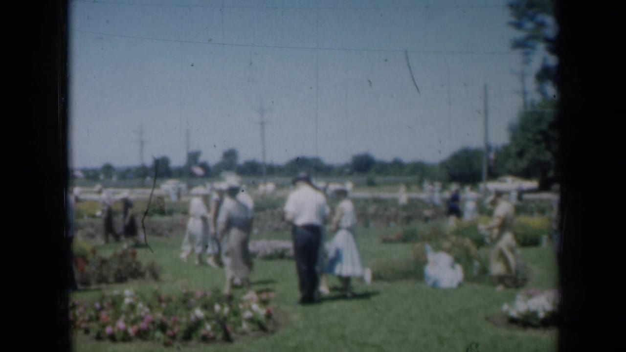 Adults walk through a sunny garden area with flower beds and lawn.