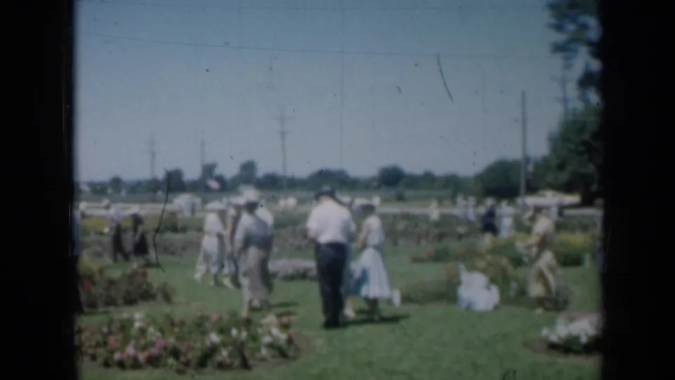 Adults in 1960s Missouri home-movie footage walk through a garden gathering with flower beds and open grass.