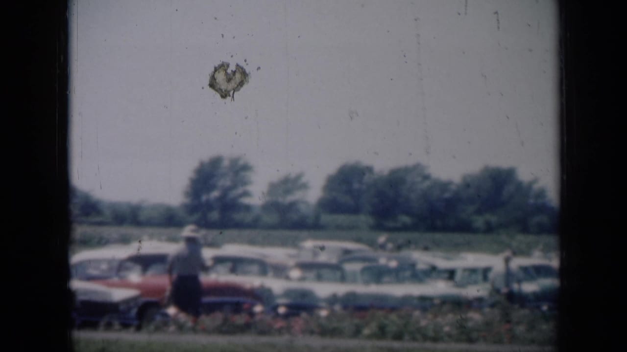 1961 Missouri home-movie frame showing people near rows of mid-century cars in a grassy outdoor area.