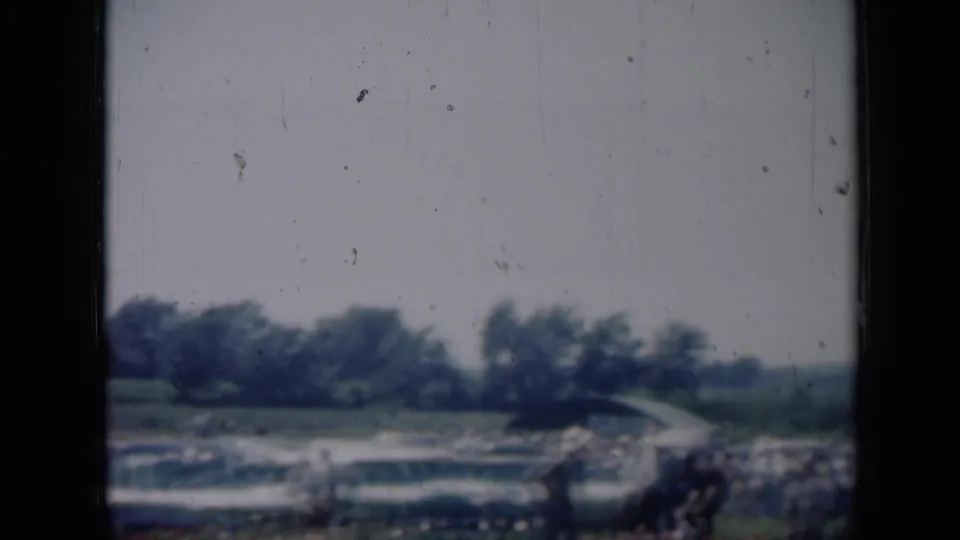 Distant view of a Missouri outdoor gathering with classic cars and people along the field edge.