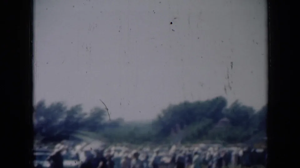 Soft 8mm frame of a large outdoor crowd in Missouri, with trees and parked cars partly visible.