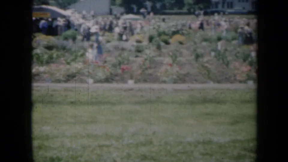 1961 Missouri 8mm frame showing distant visitors and women near planted flower beds across a lawn.