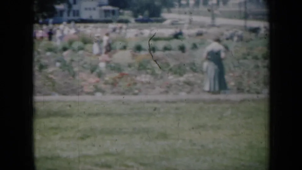 1961 Missouri garden scene with a woman in a long dress beside planted beds and groups of visitors beyond.