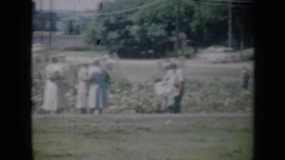 1961 Missouri home-movie frame of several women in dresses standing near flower beds in a sunny garden area.