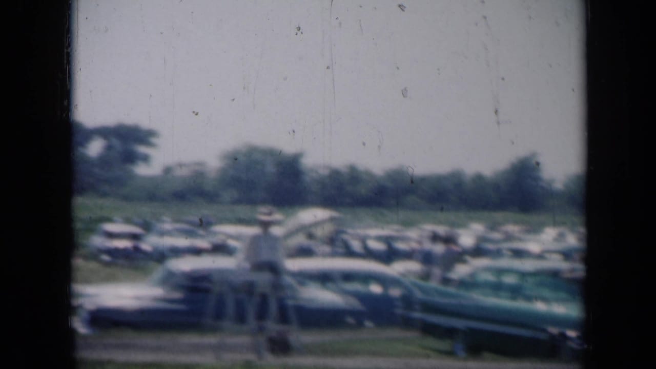 Parked cars and people in a rural field seen through dark film gate edges.