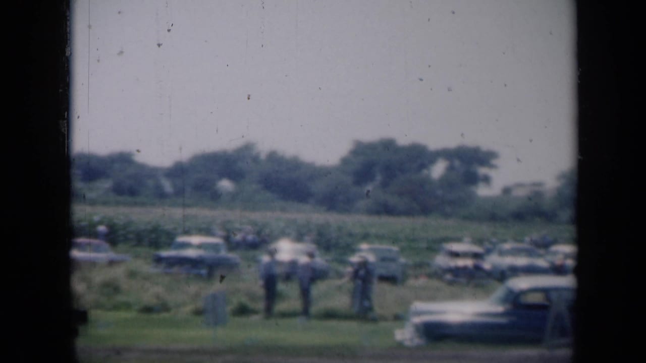 Groups of people stand near parked cars along the edge of a field.