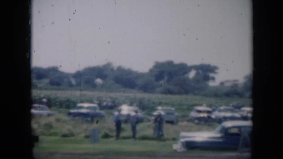 1961 Missouri scene with people in light clothing standing near rows of parked cars beside an open rural field.
