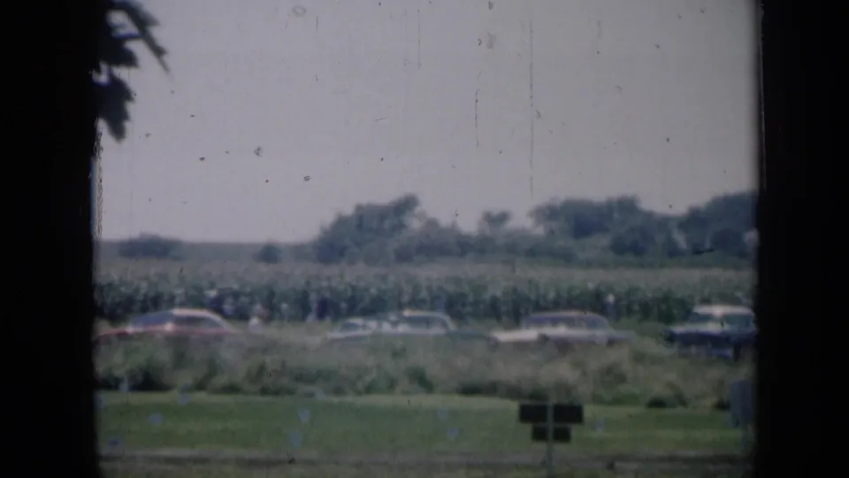 Wide 1961 Missouri home-movie view of cars parked beside crops and a tree line, with a small sign or marker in the foreground.
