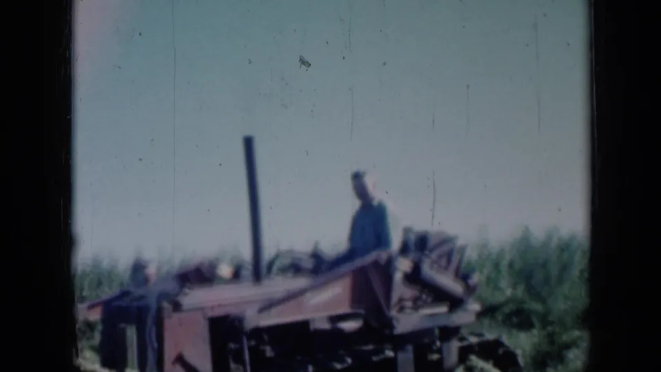 1961 Missouri home-movie frame showing a man seated on a red tractor or farm machine in a field with tall vegetation behind him.
