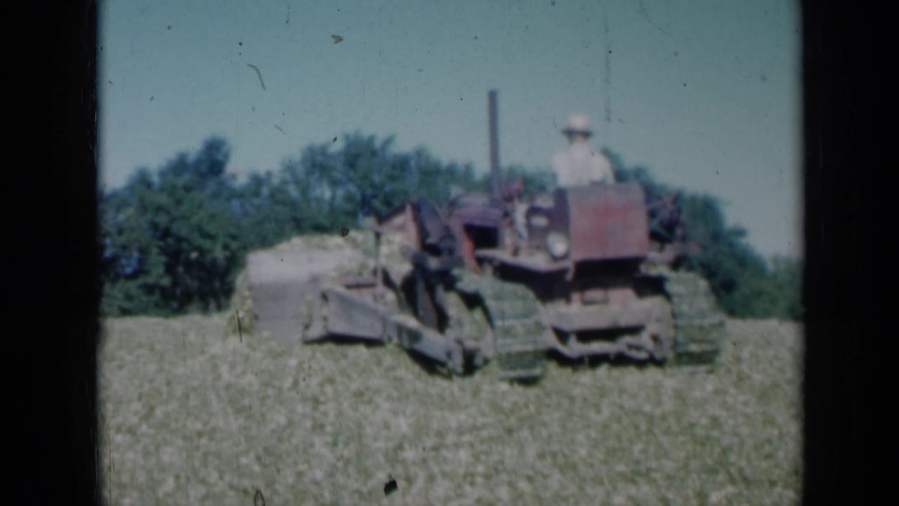 1961 Missouri 8mm frame showing a red farm tractor or crawler pushing a pile of hay or cut crop in a green field, with a seated operator and tree line behind.