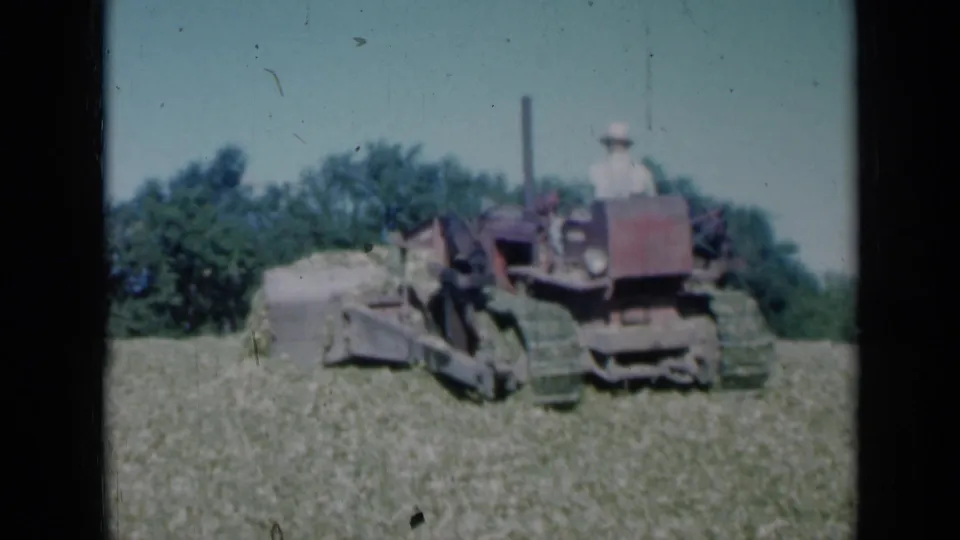 1961 Missouri 8mm frame showing a red farm tractor or crawler pushing a pile of hay or cut crop in a green field, with a seated operator and tree line behind.