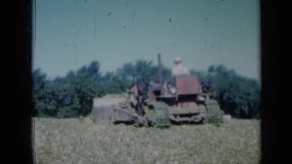 1961 Missouri home-movie frame showing the tractor slightly lower in the frame, with crop material ahead of the machine and a dark green tree line in the background.