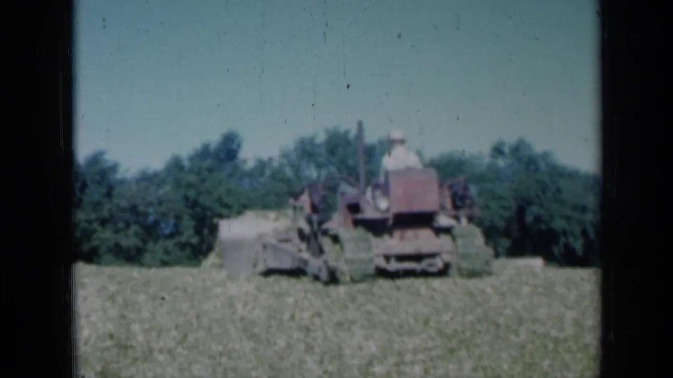 1961 Missouri 8mm frame showing the red tractor and operator in a rural field, with pushed hay or cut crop in front and trees forming the background.