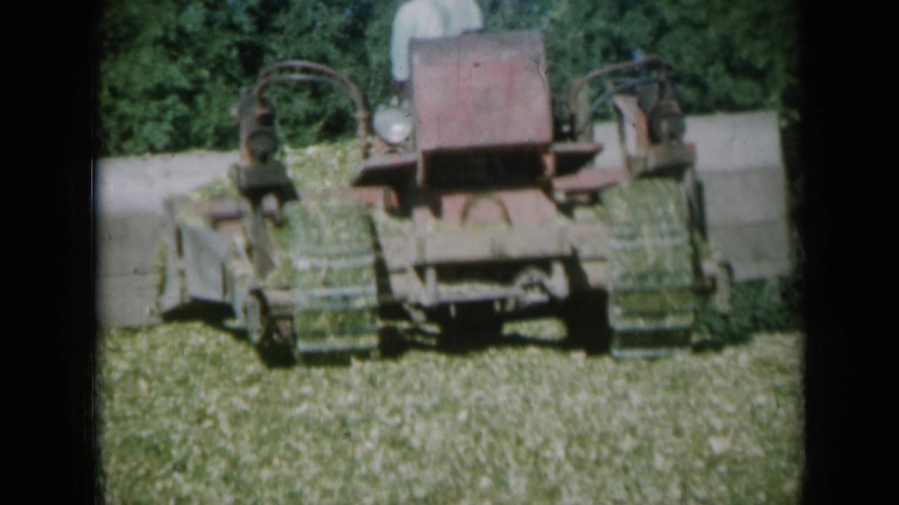 1961 Missouri farm footage showing a red tractor-mounted hay baler working in cut grass with a hat-wearing operator partly visible.