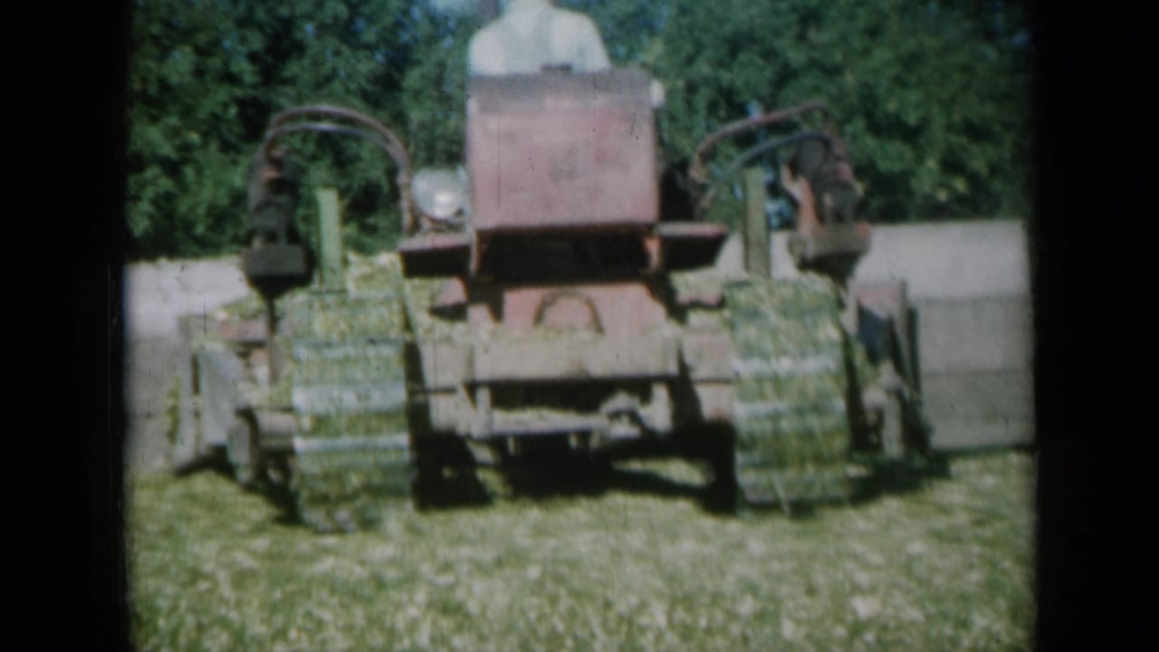 Front view of a red hay baler moving through grass with trees behind it.