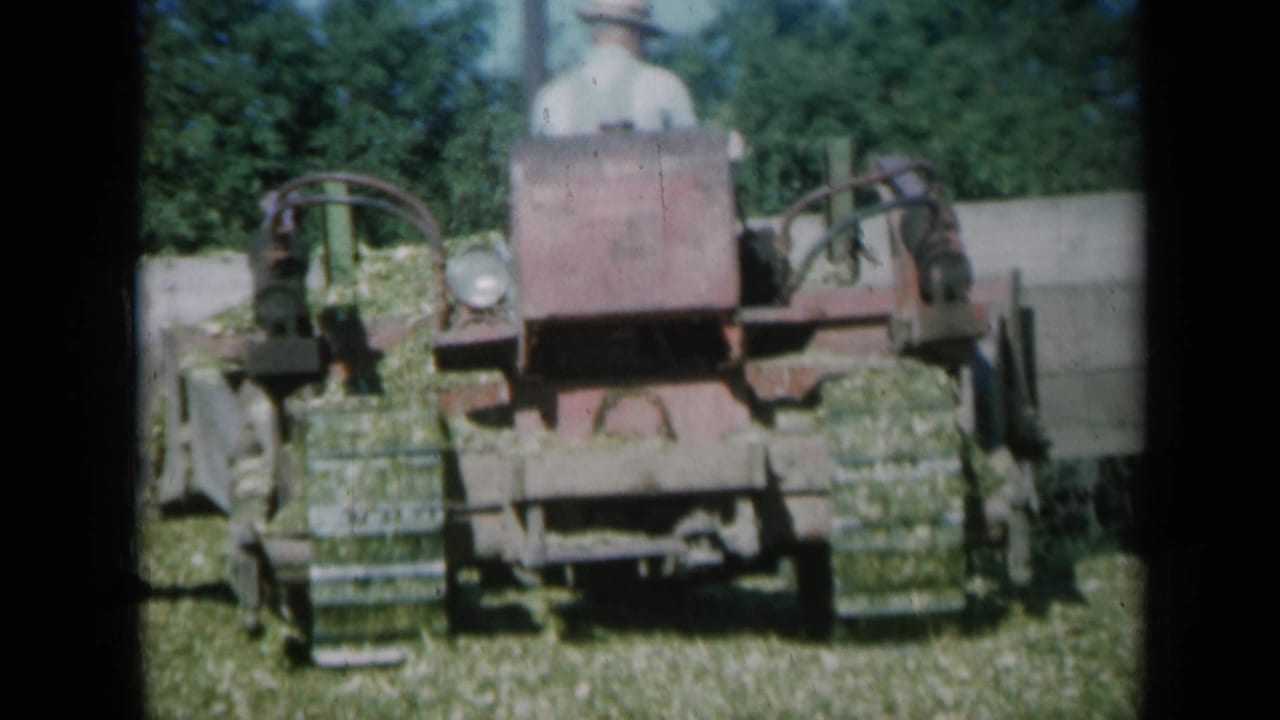 The 8mm frame shows the baler close to camera as it gathers green grass, with the operator faintly visible above the machine.