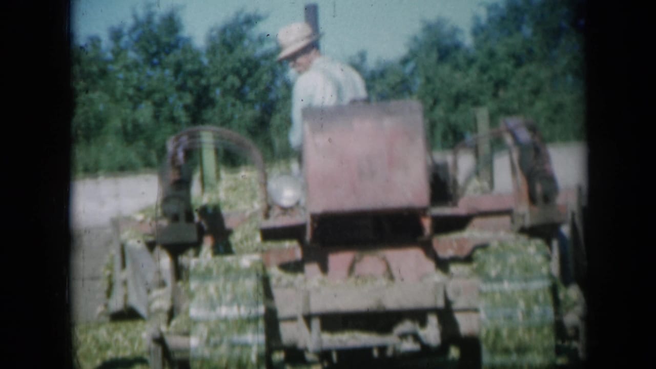 A hat-wearing operator is more visible behind the red hay baler as the machine continues across a Missouri farm field.