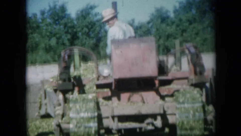 A hat-wearing operator is more visible behind the red hay baler as the machine continues across a Missouri farm field.