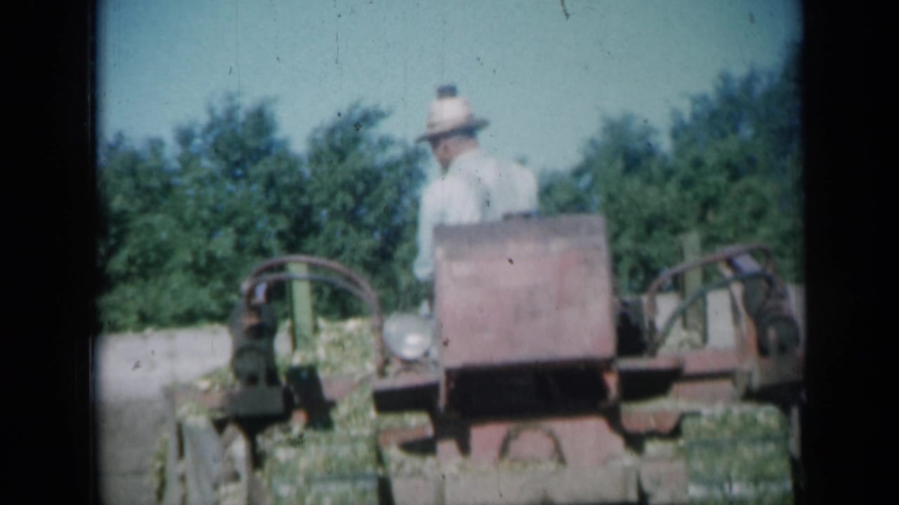 In 1961 Missouri footage, an older man in a light shirt and straw hat is seen with a red grader near trees and open ground.