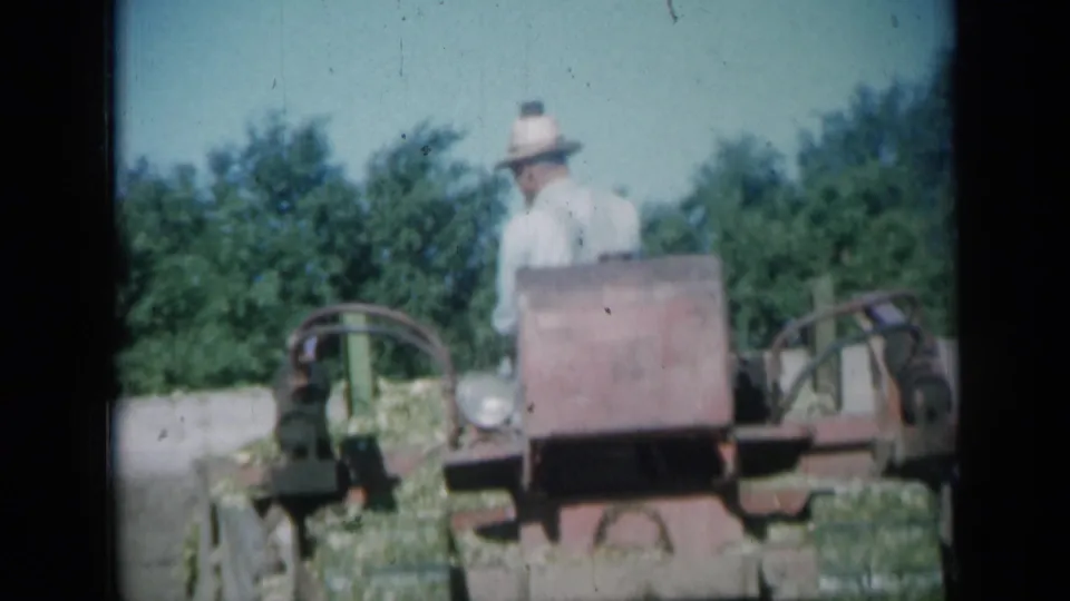 In 1961 Missouri footage, an older man in a light shirt and straw hat is seen with a red grader near trees and open ground.