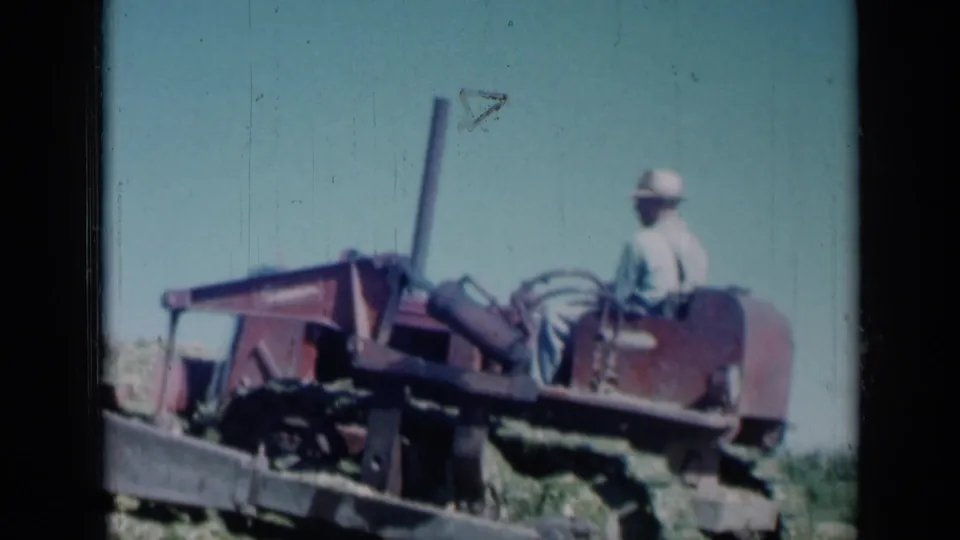 Side view of the tractor operator riding through a harvested Missouri field, with soft 8mm color and visible frame wear.
