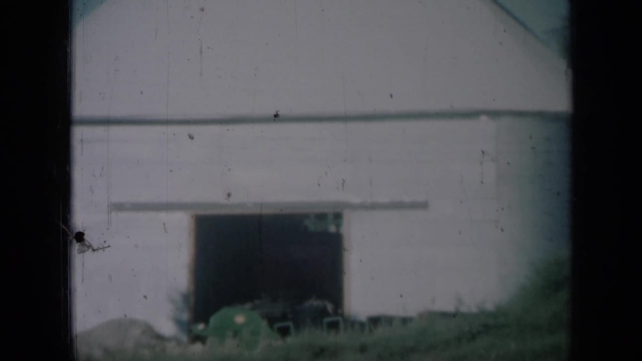 Pale farm building with an open doorway beside grass and dark film frame edges.