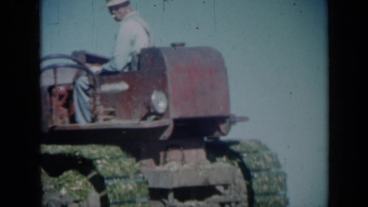 Adult operator in a light shirt drives a red tracked tractor near a farm building.