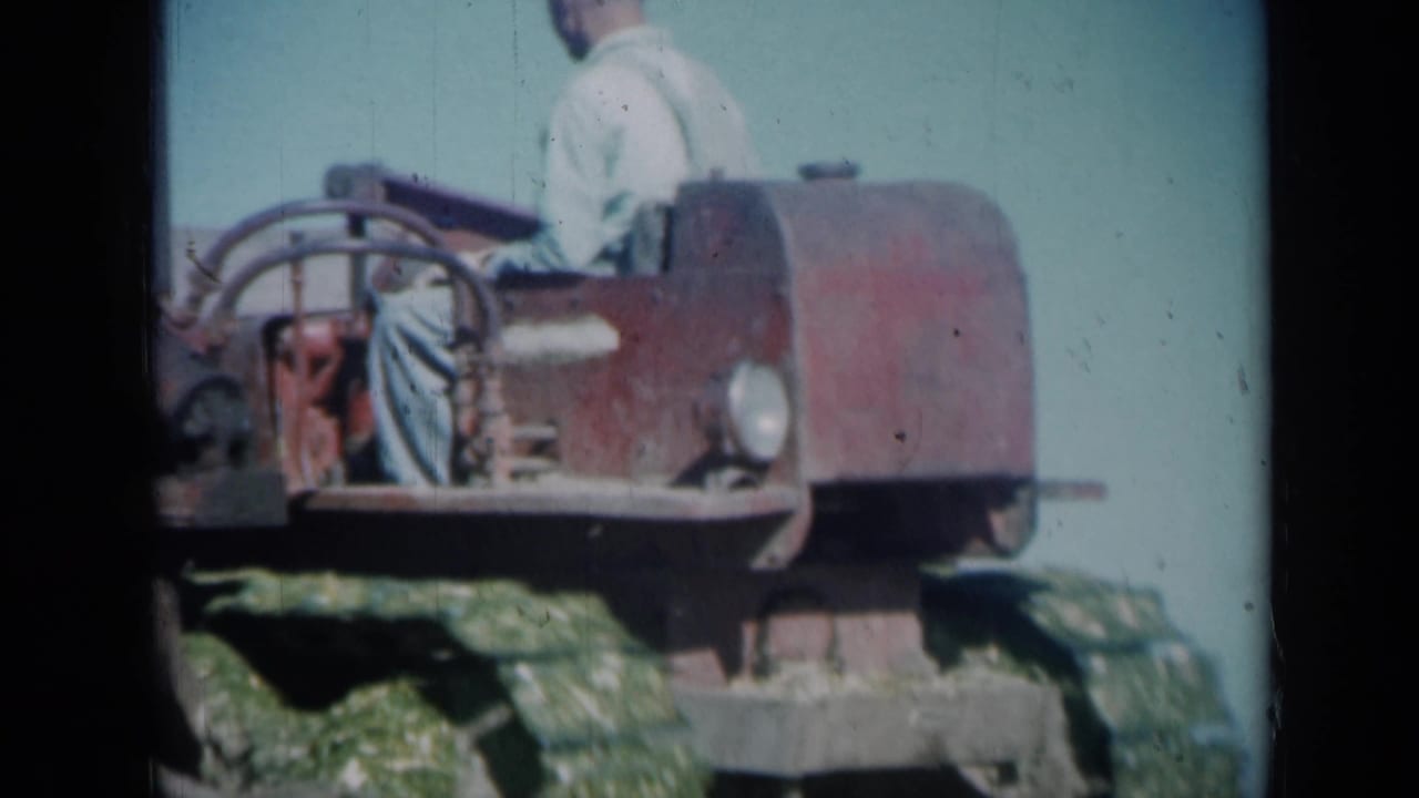 Close 1961 Missouri farm frame of an adult operator seated on a red tracked tractor, with grass and blue sky behind the machine.