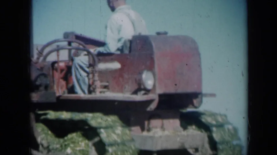 Close 1961 Missouri farm frame of an adult operator seated on a red tracked tractor, with grass and blue sky behind the machine.