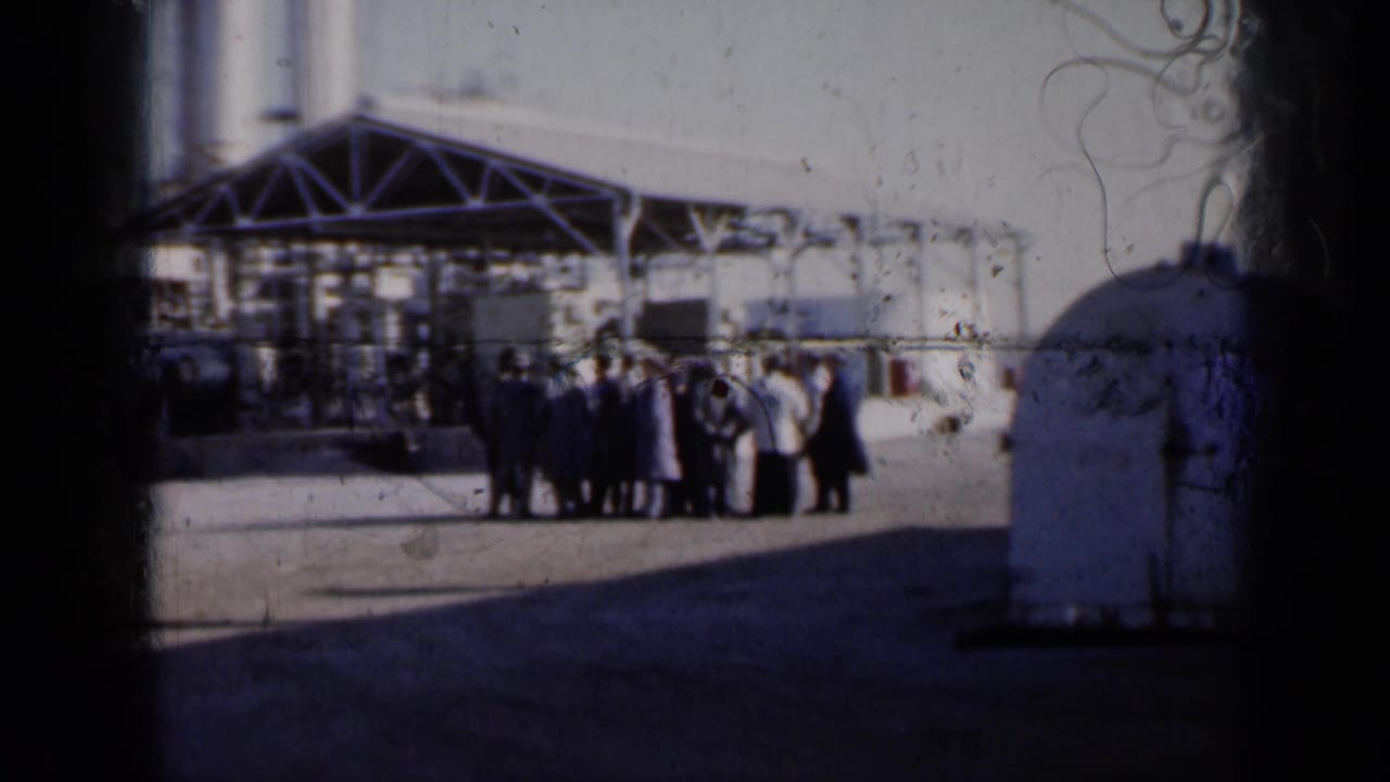 Adults gather outside a large industrial building near silos and a rounded tank.