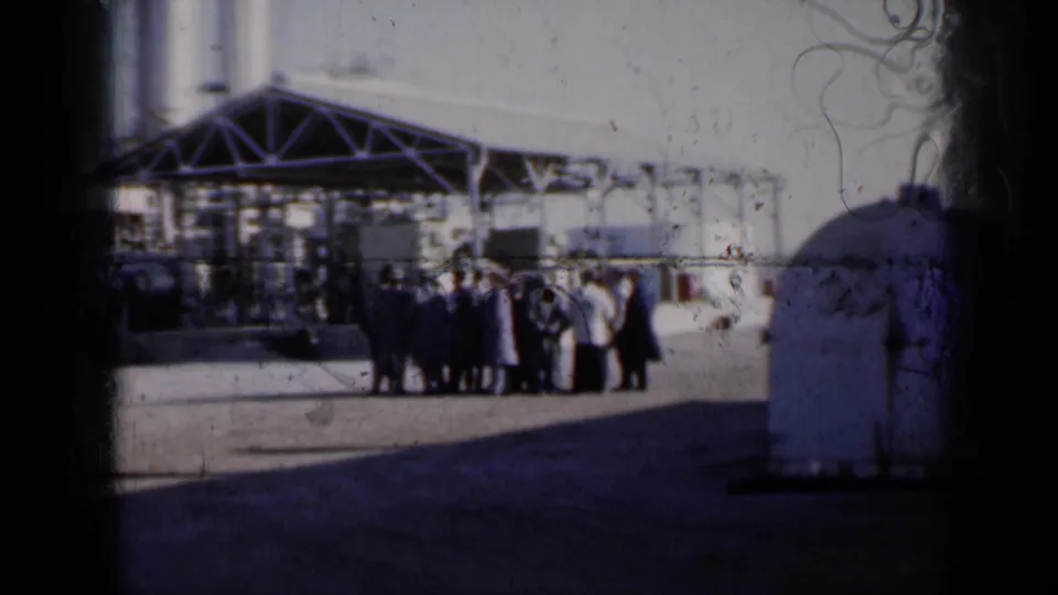 1961 Missouri 8mm frame showing adults grouped near an industrial building with silos, a metal canopy, and a rounded tank.